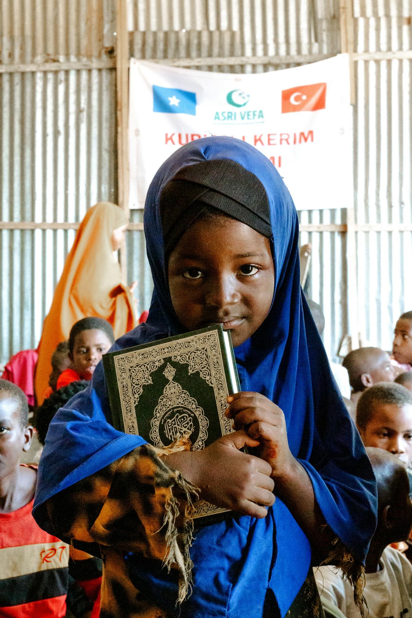 A young girl in a blue hijab holding the Quran at a community event indoors.